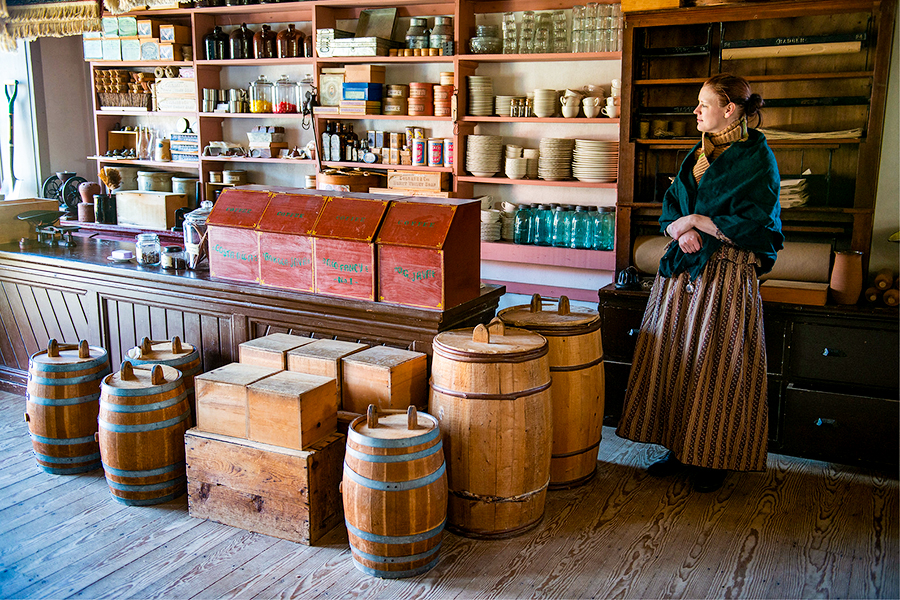 a woman in clothes from the time stands the the side of a store counter that is decorated to match the time period of the woman