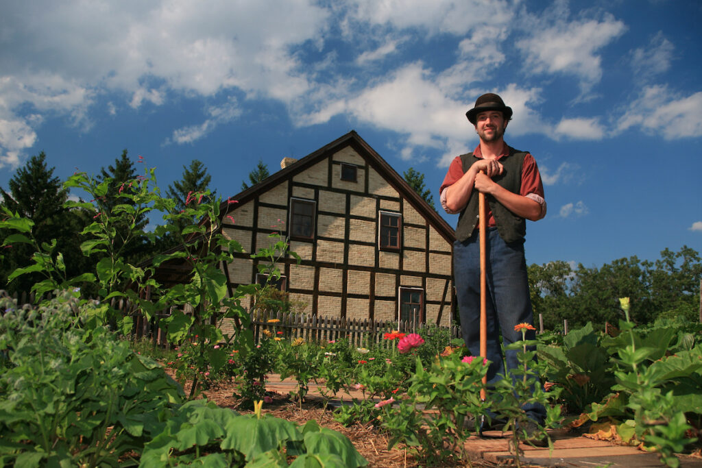 A man stands leaning lightly on his garden hoe in a flower garden that he tends as part of his employment at Old World Wisconsin. 