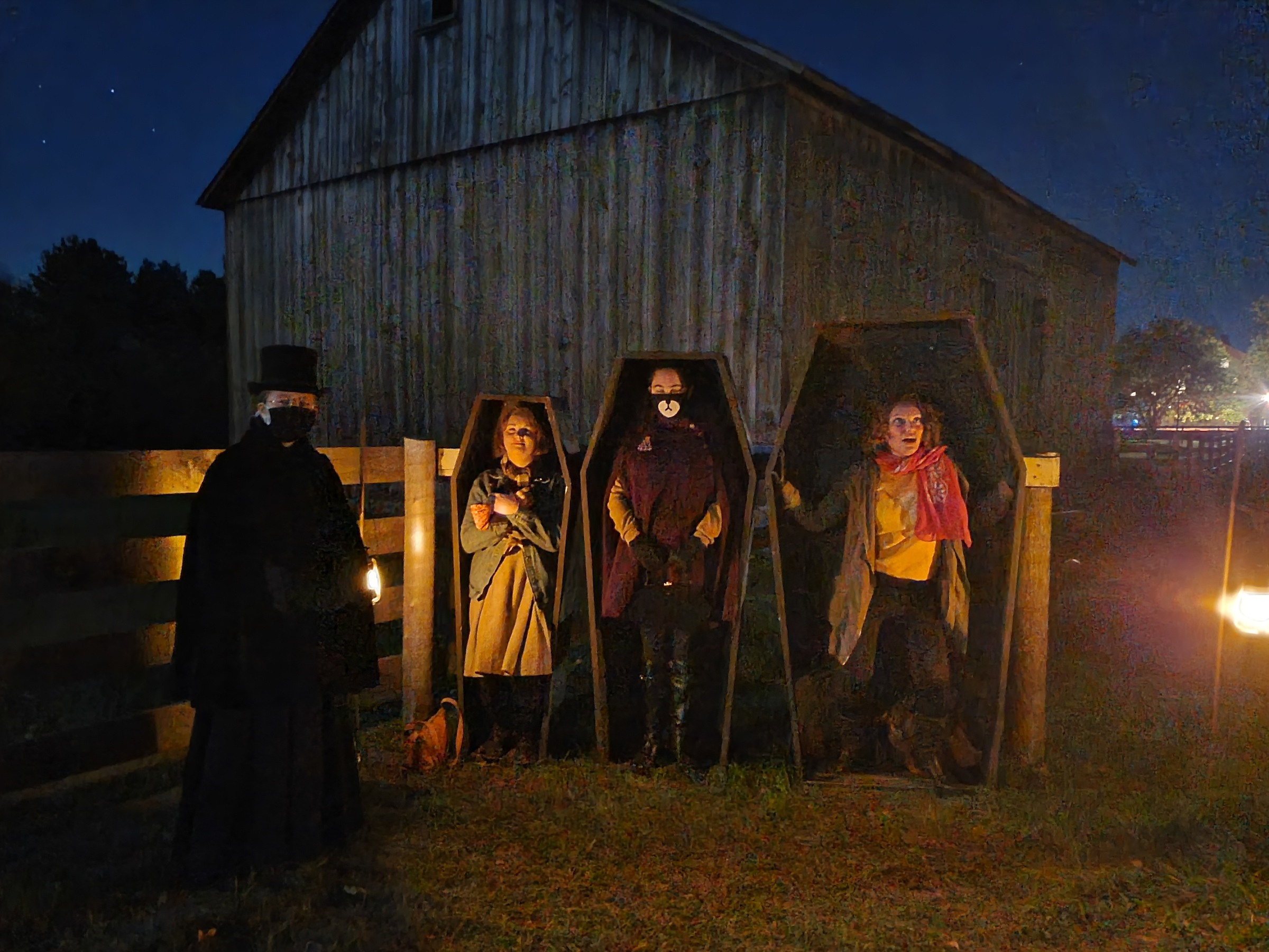 Three people, in costumes, standing in coffins outside during the night, with a person dressed in all black beside them