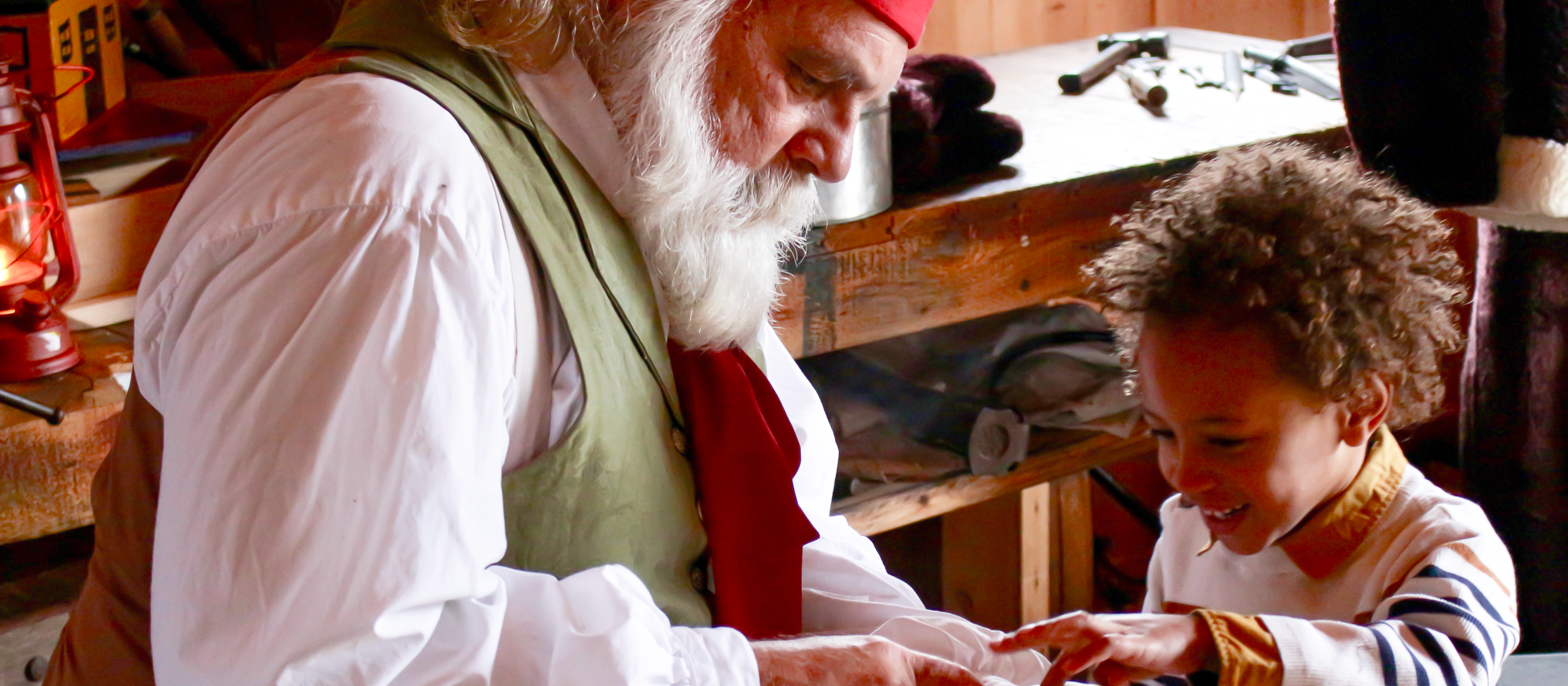 An image of a man, with a white beard, wearing a white collared shirt with a green vest and wearing a red hat, dressed up as Santa looking at something with a young boy.
