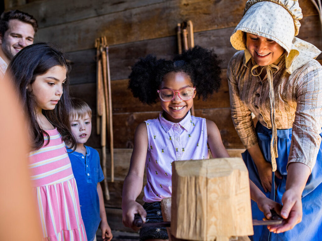 A young black girl with hair poms smiles excitedly as she's taught how to work with wood by a white woman in period clothing, as some children and a man look on with curiosity