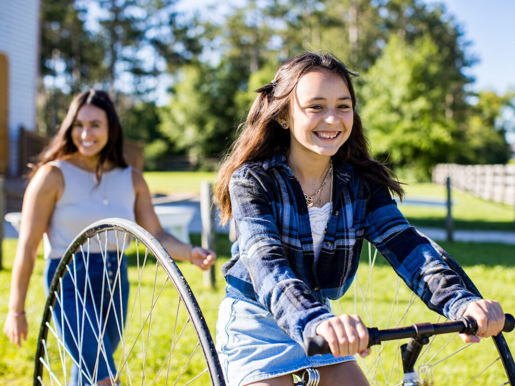A young girl riding a bike, smiling, with a woman following behind her in the background