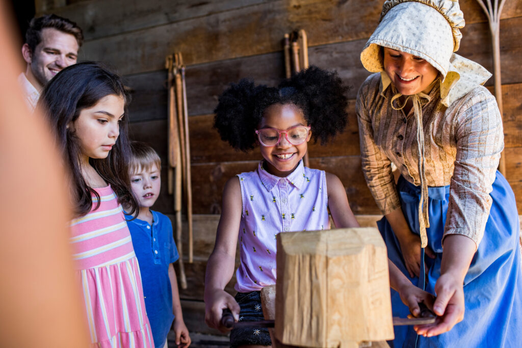 A young black girl with cute hair poms smiles excitedly as she's taught how to work with wood by a white woman in period clothing, as some children and a man look on with curiosity. OWW Summer 2022 Photoshoot