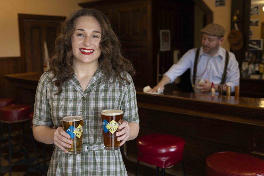 A woman bartender smiling at the camera with two glasses of beer in each hand. The glasses are printed with a yellow and blue logo and says "Wittnebel's Tavern" while a male bartender cleans behind her