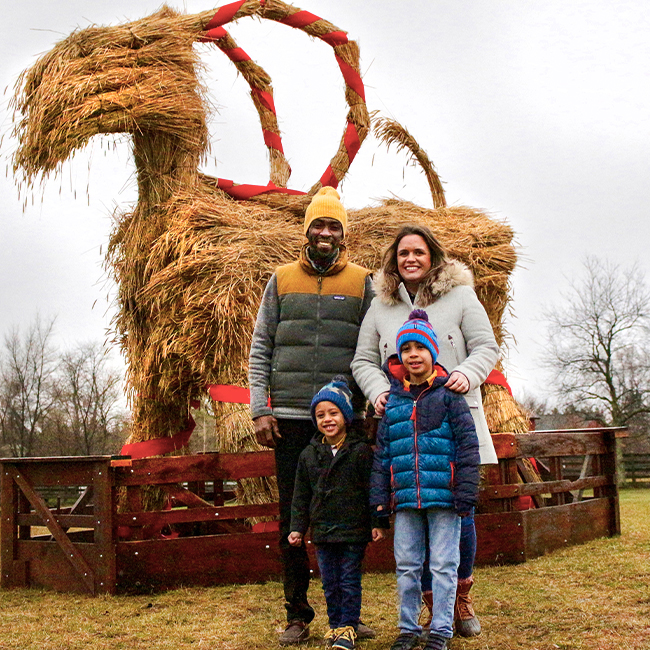 A family photo in front of the Julblock, a straw goat. The family, a black man wearing a yellow hat with a yellow and grey jacket standing next to a white woman with brown hair, wearing a light grey coat. There are two children standing in front, a small boy wearing a blue and red hat with a blue jacket and blue jeans and a smaller boy wearing a blue hat and dark jacket and dark jeans. 